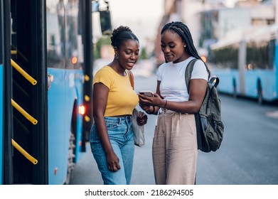 African american female friends waiting for a bus while at a bus stop and using a smartphone. Riding, sightseeing, traveling to work, city tour, togetherness. Bus schedule app, browsing social media. - Powered by Shutterstock