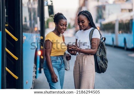 African american female friends waiting for a bus while at a bus stop and using a smartphone. Riding, sightseeing, traveling to work, city tour, togetherness. Bus schedule app, browsing social media.