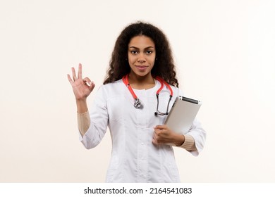 African American Female General Doctor with tablet PC. Data on tablet. Digital Medical App. Physician in Uniform with Stethoscope Shows Ok sign on Light Background