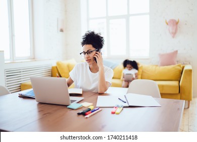 African American female in glasses speaking on mobile phone while using laptop for remote work taking care of daughter at home 
