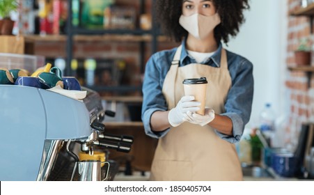 African American Female Hipster Waitress In Face Mask And Gloves Holds Takeaway Drink Order In Hands Giving Cup In Cafe. Coffee Shop Worker Offering Takeout Safe Restaurant Coffee Away Delivery