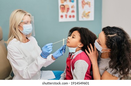 African American Female Kid Having PCR Test During Coronavirus Epidemic. Doctor Wearing A Protective Shield Using A Cotton Swab Makes A Kid's Nasal Cavity Test For Covid 19 Virus