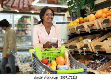 African American Female Lady Holding Cellphone Doing Grocery Shopping Standing With Shop Cart Smiling To Camera In Modern Supermarket Indoor. Customer With Smartphone Buying Food Posing In Store