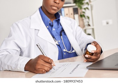 African American Female Medic Doctor Gp Therapist Wear White Coat Holding Pills Bottle In Hand Writing Medical Prescription Sitting At Work Desk Prescribing Pharmacy Medicine Concept. Closeup View