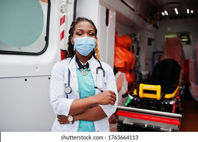 African american female paramedic in face protective medical mask standing in front of ambulance car.