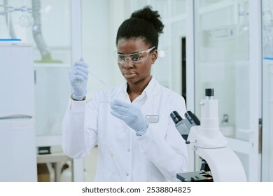 African American female scientist holding pipette, wearing safety goggles and lab coat, focusing intently on experiment in a well-equipped, brightly lit laboratory - Powered by Shutterstock