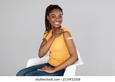 African American Female Smiling After Receiving Coronavirus Vaccine Shot On Grey Studio Background. Millennial Black Woman Getting Vaccinated Against Covid, Showing Arm With Bandage