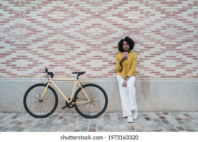 African American Female standing near parked yellow fixie bike and messaging on social media on smartphone on a pink brick wall