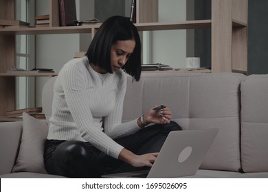 African American Female With Straith Short Hair In Casual Clothes Seating On Couch And Entering Credit Card Credentials On Laptop While Shopping Online At Home