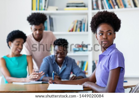 African american female student learning at desk at school with teacher and group of students at classroom