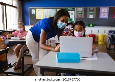 African american female teacher wearing face mask teaching a girl to use laptop at elementary school. back to school and education concept - Powered by Shutterstock