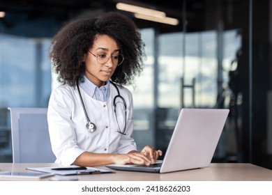 African american female therapist dressed in uniform typing on laptop while sitting at workplace in hospital. Pretty physician using modern device and internet for helping and supporting patients. - Powered by Shutterstock