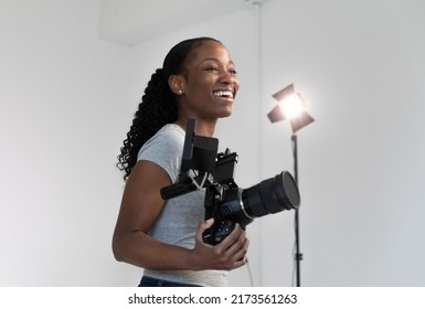 African American Female Videographer Posing with Video Camera in Hand on Film Set. Camera Woman is Proud and Smiling Looking Off Camera.