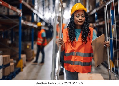 African american female warehouse employee, wearing safety gear, navigates a cart loaded with boxes down an aisle. Focus, safety, and effective inventory management in a workspace. - Powered by Shutterstock