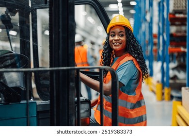 African american female warehouse worker wearing an orange vest and hard hat smiles cheerfully beside a forklift in the industrial setting, emphasizing a positive and safe atmosphere. - Powered by Shutterstock