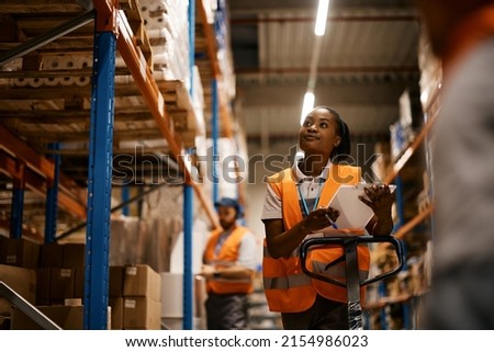 African American female worker going through inventory list while checking stock at distribution warehouse.