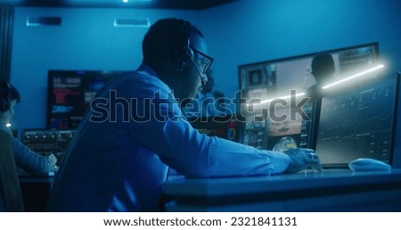 African American flight control employee in headset monitors space mission on multi-monitor computer in command center. Team clap hands after successful space rocket launch displayed on big screens.