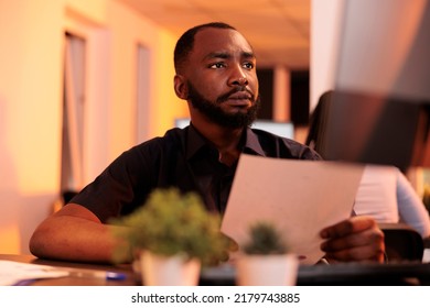 African American Freelancer Looking At Research Documents To Plan Solutions For Paperwork Report, Using Computer In Office. Analyzing Corporate Information On Files During Sunset.