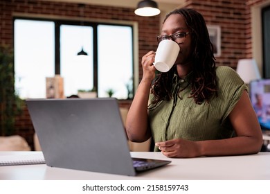 African American Freelancer Taking A Break From Remote Work Drinking Coffee Or Tea At Desk With Laptop Looking At Screen. Student At Home Watching Video Content On Portable Computer In Living Room.