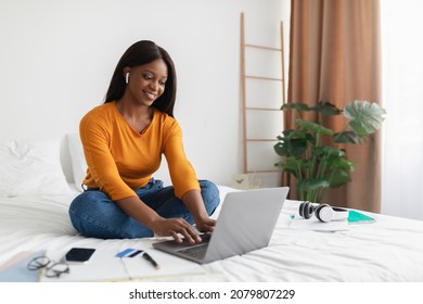 African American Freelancer Woman Using Laptop Wearing Earbuds Typing On Computer Working Online In Bedroom At Home. Freelancer Lady Browsing Internet Sitting On Bed Indoor. Technology And Freelance