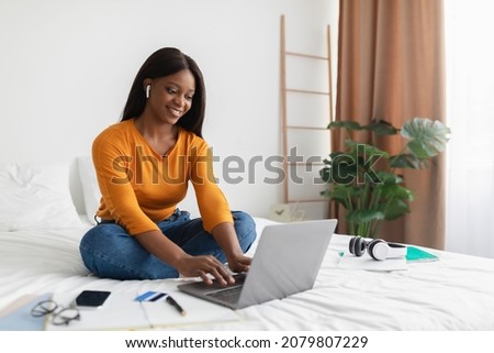African American Freelancer Woman Using Laptop Wearing Earbuds Typing On Computer Working Online In Bedroom At Home. Freelancer Lady Browsing Internet Sitting On Bed Indoor. Technology And Freelance
