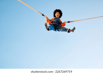 An african american girl child with afro curly hair jumping on a trampoline with insurance elastic bands in an amusement park and carousels.