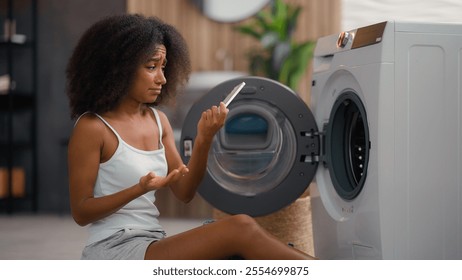 African American girl ethnic lady biracial woman housewife home laundry clothes open washing machine housekeeping take clean wet jeans pants find mobile phone in pocket broken smartphone shock upset - Powered by Shutterstock