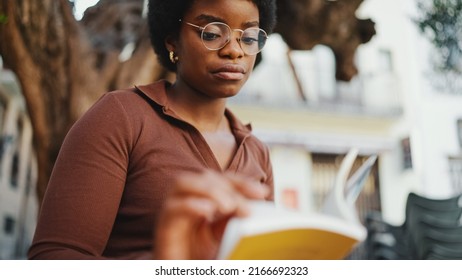 African American Girl In Glasses Looking Serious Reading Book Outdoors. Female Student Searching For Information In Textbook. University Campus Life