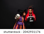 african american girl in halloween costume holding skull and blowing on witch cauldron with potion near brother isolated on black