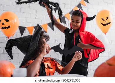 African American Girl In Halloween Costume Holding Paper Cut Bat Near Happy Brother With Toy Spider Showing Thumb Up