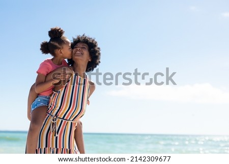 African american girl kissing mother while enjoying piggyback ride on her at beach against blue sky. unaltered, family, lifestyle, togetherness, enjoyment and holiday concept.