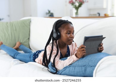 African american girl lying on couch, listening to music and using tablet at home, copy space. Childhood, expression, technology, communication and domestic life, unaltered. - Powered by Shutterstock