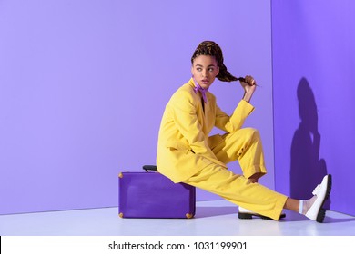 african american girl posing in yellow suit sitting on purple suitcase, on trendy ultra violet background