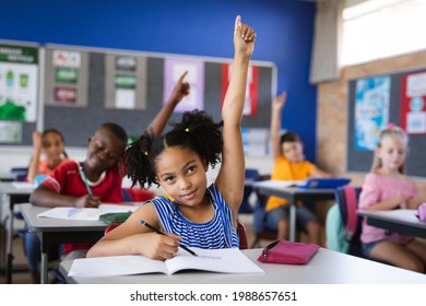 African American Girl Raising Her Hands While Sitting On Her Desk In The Class At School. School And Education Concept