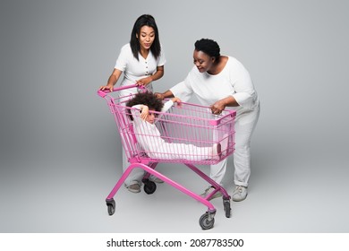 African American Girl Sitting In Shopping Trolley Near Happy Mom And Granny On Grey