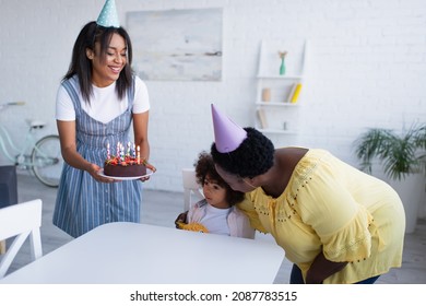 African American Girl Sitting At Table Near Granny Embracing Her And Mother Holding Birthday Cake