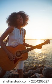 African American Girl Standing By The Ocean At Sunset Playing Guitar Enjoying Freedom Outdoors On Relaxing Beach Vacation