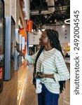 African American girl Using Self-Service Kiosk at Fast Food Restaurant. Black Woman in striped shirt interacting with a self-service kiosk in a store, holding a smartphone and headphones. 