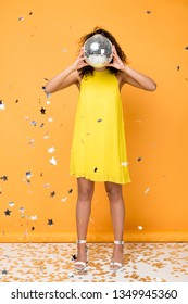 African American Girl In Yellow Dress Covering Face With Disco Ball Near Shiny Confetti Stars On Orange 