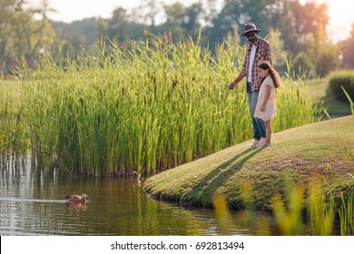 African American Granddaughter And Her Grandfather Feeding Duck On Lake In Park 