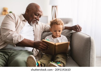 African American Grandfather And Little Grandson Reading A Book Sitting Together On Sofa At Home. Senior Male Teaching His Grandchild To Read On Weekend. Family Moments Concept