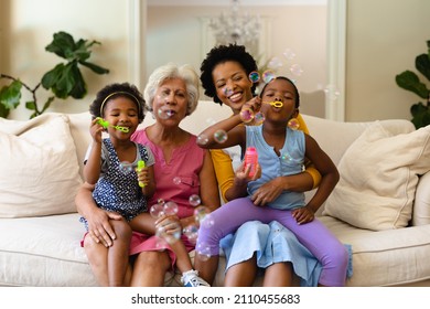 African american grandmother, mother and two granddaughters blowing bubbles sitting on couch. family, love and togetherness concept, unaltered.