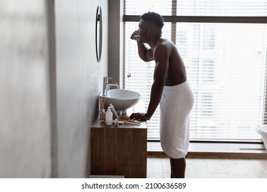 African American Guy Brushing Teeth With Toothbrush Standing Wrapped In Towel Near Mirror And Basin In Modern Bathroom At Home. Beauty Care Concept. Side View, Full Length Shot