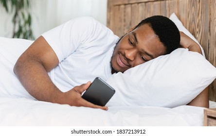 African American Guy With Cellphone Lying In Bed Using Mobile Phone Application At Home. Guy Looking At Smartphone Waiting For Call Resting In Bedroom