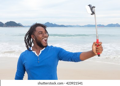 African American Guy With Dreadlocks At Beach Taking Picture With Selfie Stick With Ocean And Sky In The Background