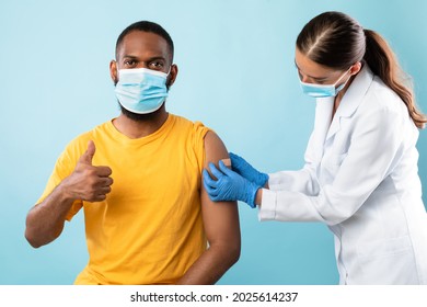 African American Guy In Face Mask Showing Thumb Up Gesture After Covid Vaccine Shot, Recommending Anti Coronavirus Vaccination, Female Doctor Putting On Band Aid Over His Arm, Blue Studio Background