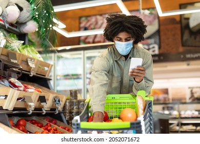 African American Guy In Face Mask Doing Grocery Shopping Using Cellphone Walking With Shop Cart In Supermarket. Customer Buying Food Products On Weekend. Selective Focus