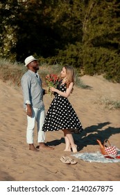 African American Guy Gives A Big Bouquet Of Flowers To His Girlfriend. A Young Interracial Couple Had A Picnic On The Beach. Romantic Date On A Summer Day 