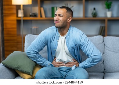 African american guy having stomach ache after eating touching aching stomach suffering from pain sitting on sofa at home. Food poisoning, gastritis, abdominal inflammation and appendicitis. - Shutterstock ID 2244844159