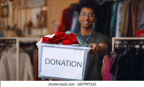 African American Guy Holding Donation Box And Looking At Camera Posing In Charity Shop. Portrait Of Smiling Afro Man Volunteer Working In Volunteering Organization And Sorting Clothes For Donation